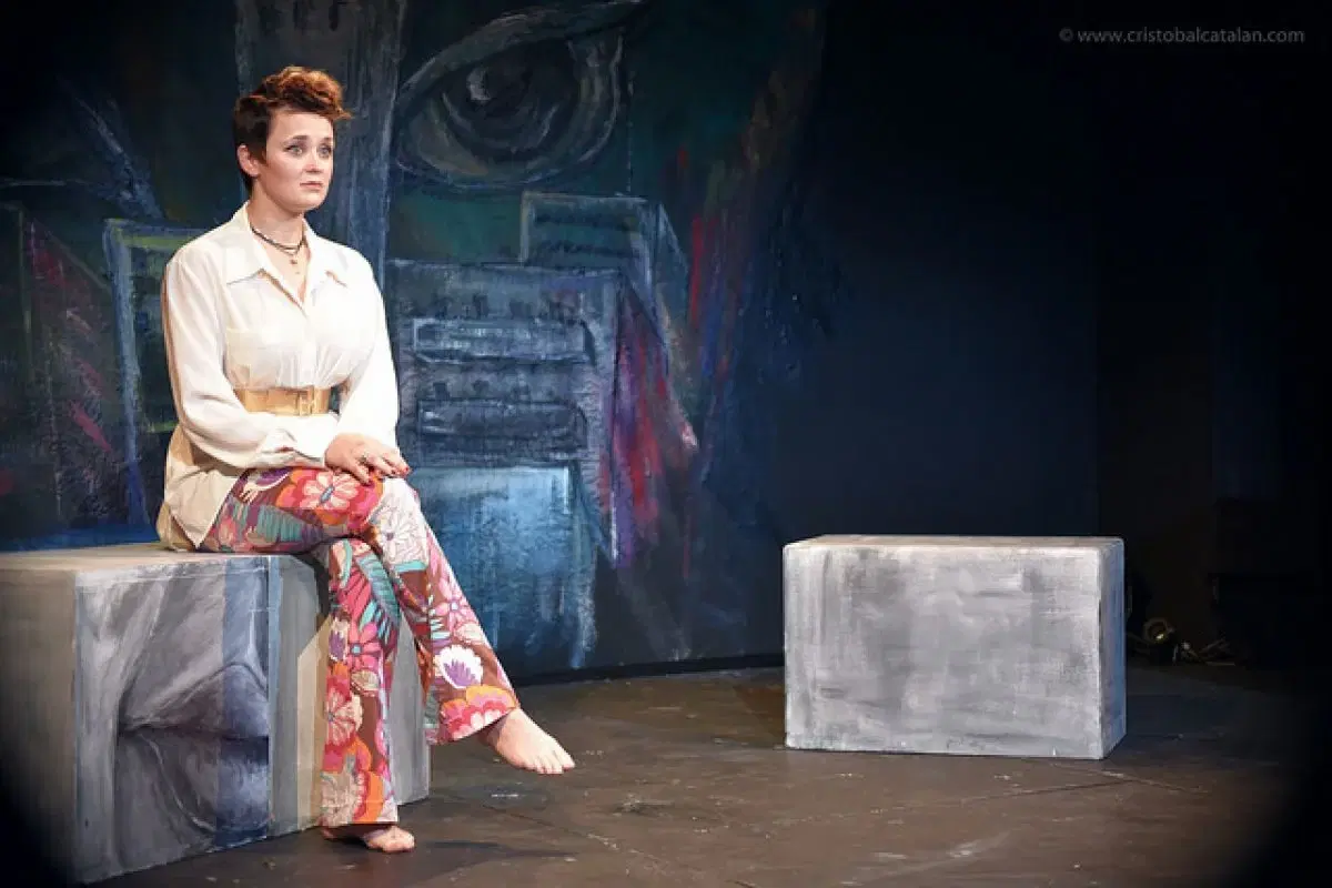 A pensive actress on stage, sitting on a metallic cube, with colorful trousers, set against a backdrop featuring an abstract face painting.