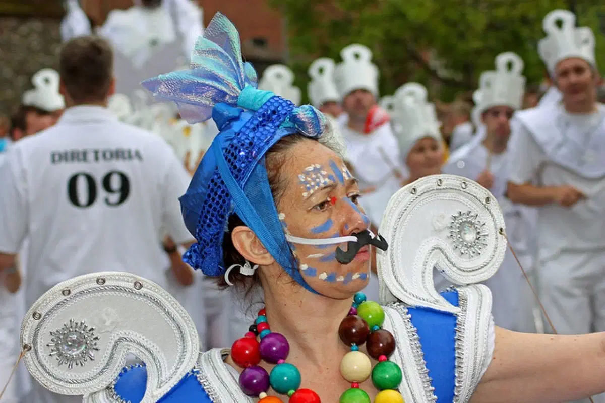 A colorful parade participant with face paint and a vibrant headpiece marches amidst a group dressed in white with chef hats.