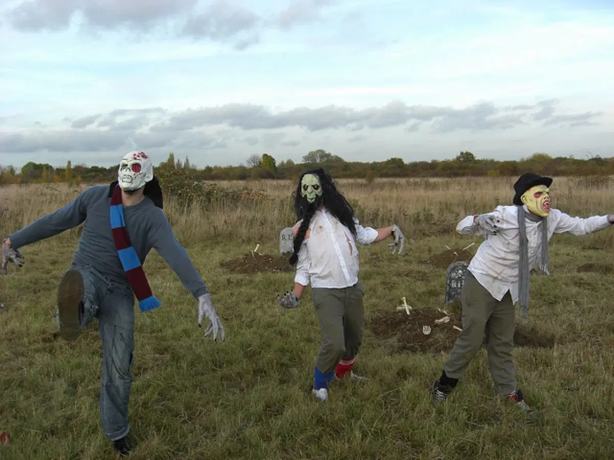 Three individuals dressed as zombies, with theatrical makeup and costumes, are pretending to stagger through a field in a classic undead pose.