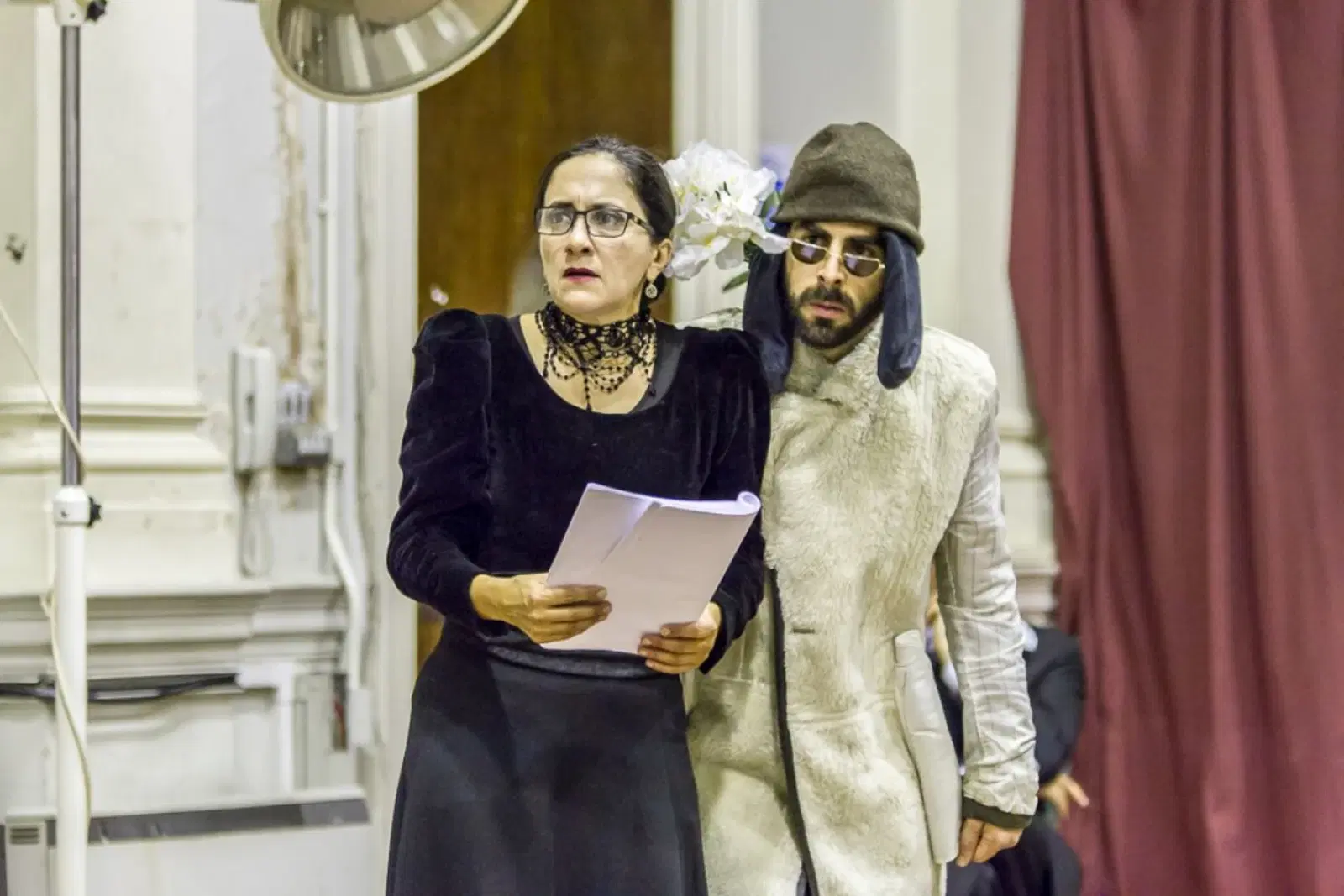 A duo performing a dramatic reading or act on stage, with the woman dressed in black and holding papers, alongside a man in a quirky hat and sunglasses, seemingly immersed in their roles.