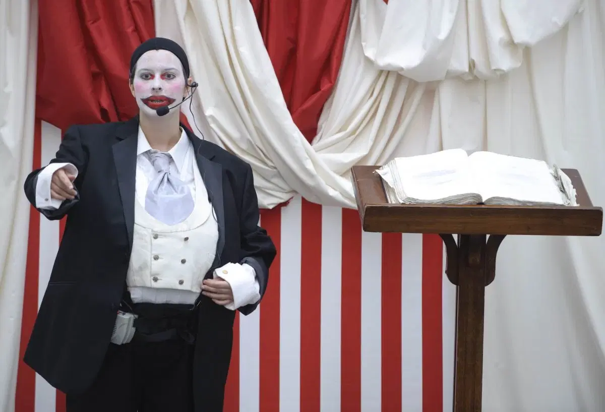 Person in mime attire performing on stage with a book on a stand, against a red and white striped backdrop.