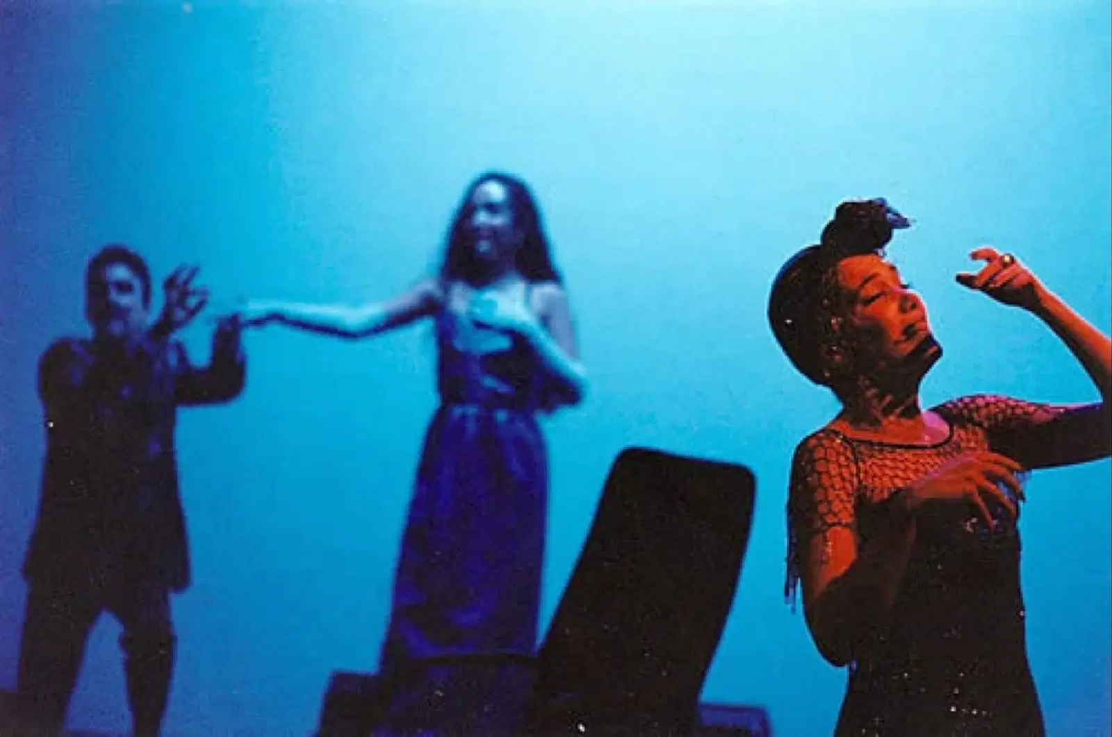 Three performers engaged in a dramatic moment on stage, bathed in moody blue lighting, with one in the foreground capturing attention through expressive gesturing.