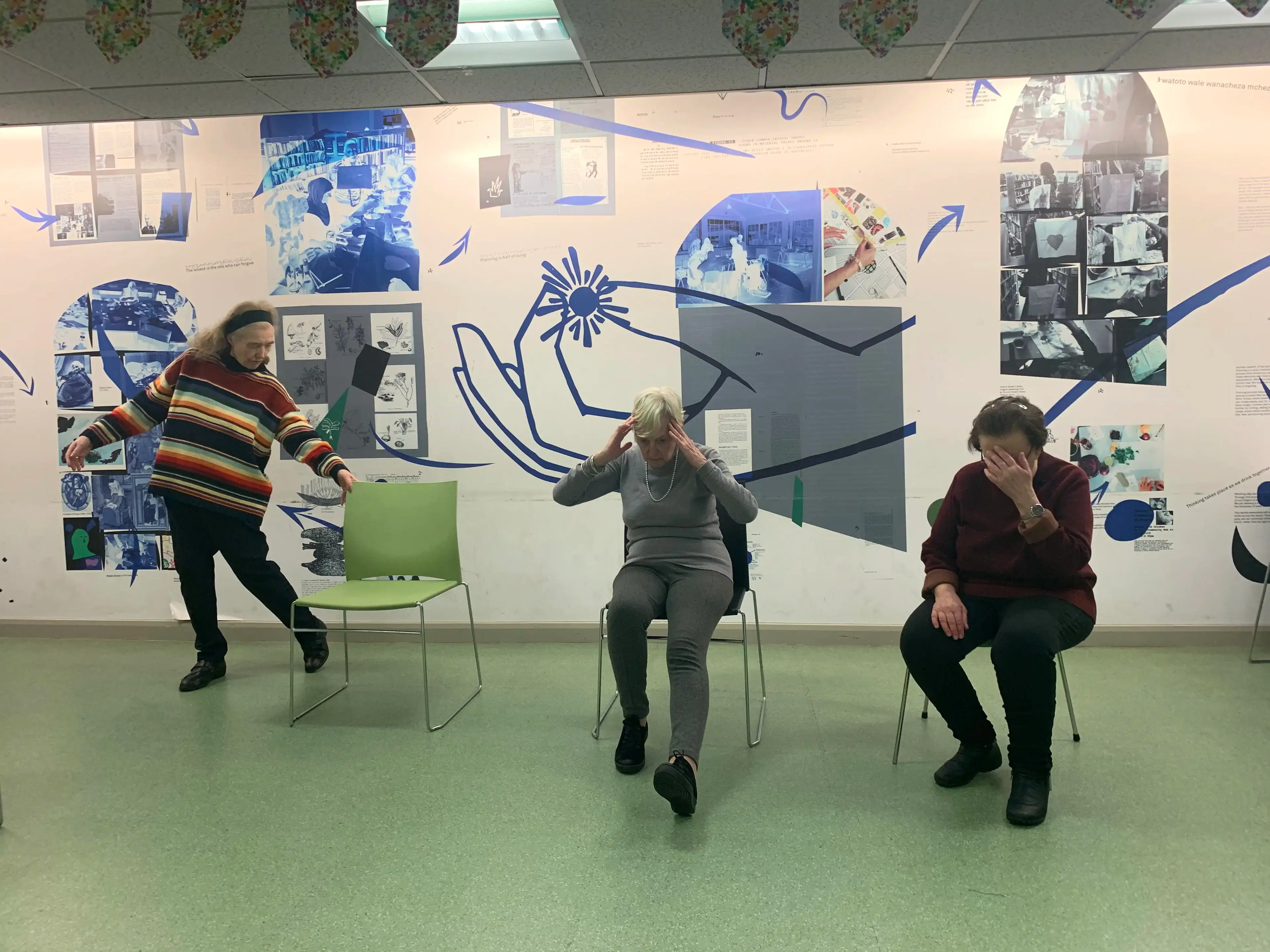 Three people in a room with illustrated walls experiencing a moment of playful chaos, as one person seems to be dancing, the middle one covers her eyes in jest, and the third looks amused, with chairs and a table suggesting a casual meeting or activity space.