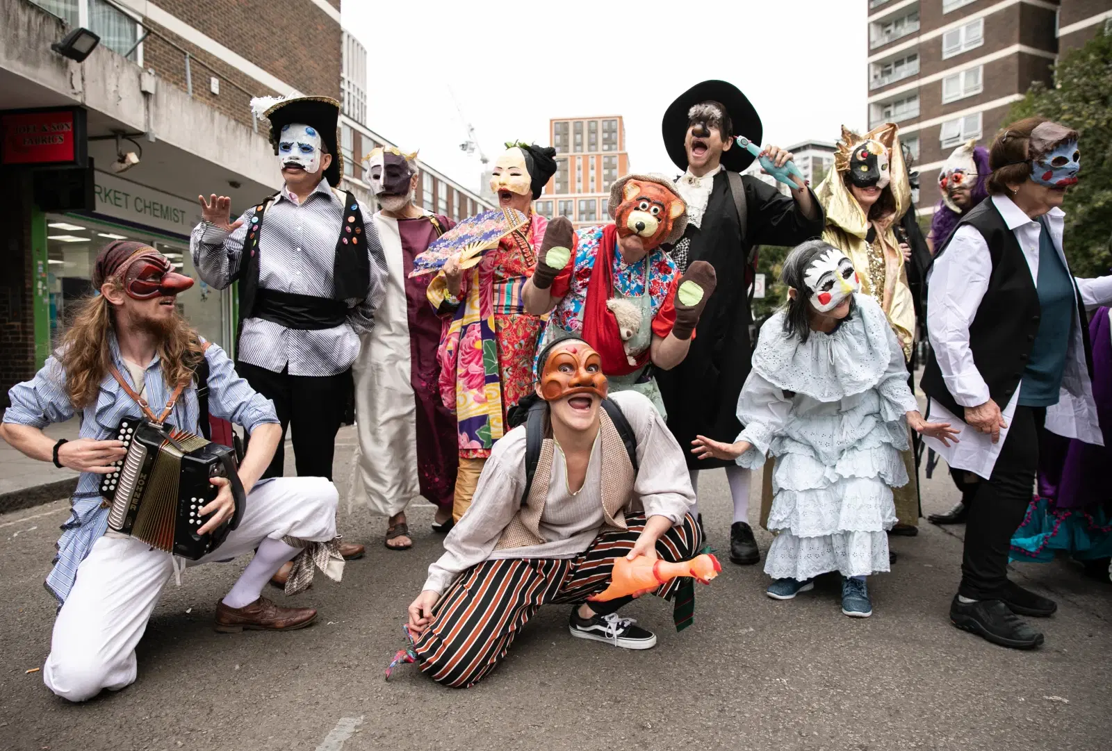 A group of individuals dressed in vibrant, eclectic costumes and masks pose theatrically on a city street. One person kneels at the front playing an accordion, while others strike expressive, playful poses. Urban buildings and shops are visible in the background.