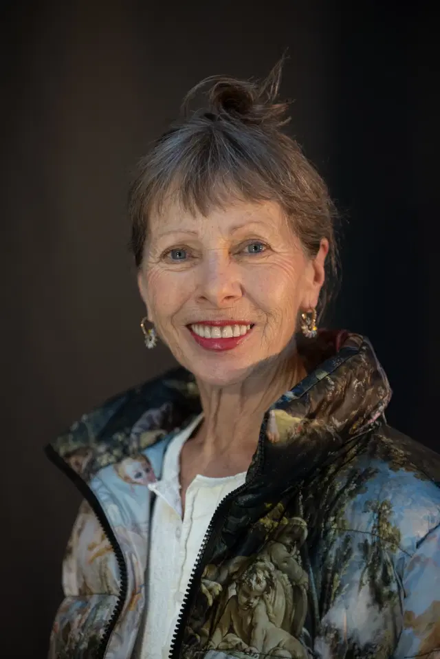 A smiling older woman with short gray hair and light blue eyes. She is wearing a patterned puffer jacket and a white shirt, along with gold hoop earrings. The background is dark, drawing attention to her bright expression.