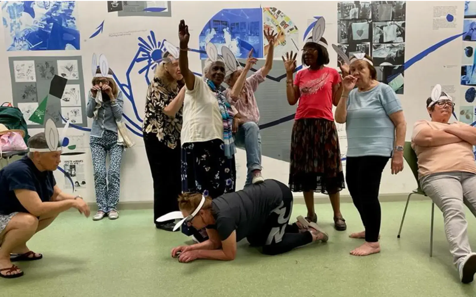 A group of people expressing themselves actively in a playful and theatrical setting, possibly during a workshop or a group activity, with some wearing whimsical hats and one person playfully kneeling on the floor.