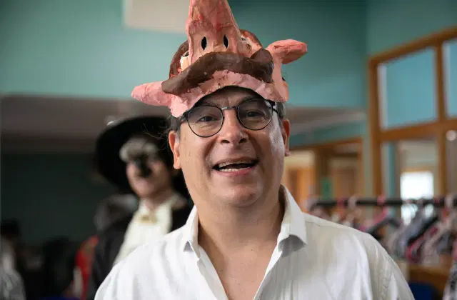 A man wearing glasses and a white shirt is smiling at the camera. He is wearing a pink, rudimentary, animal-like mask on his head. In the background, another person, slightly out of focus, is dressed in a costume and a black hat.